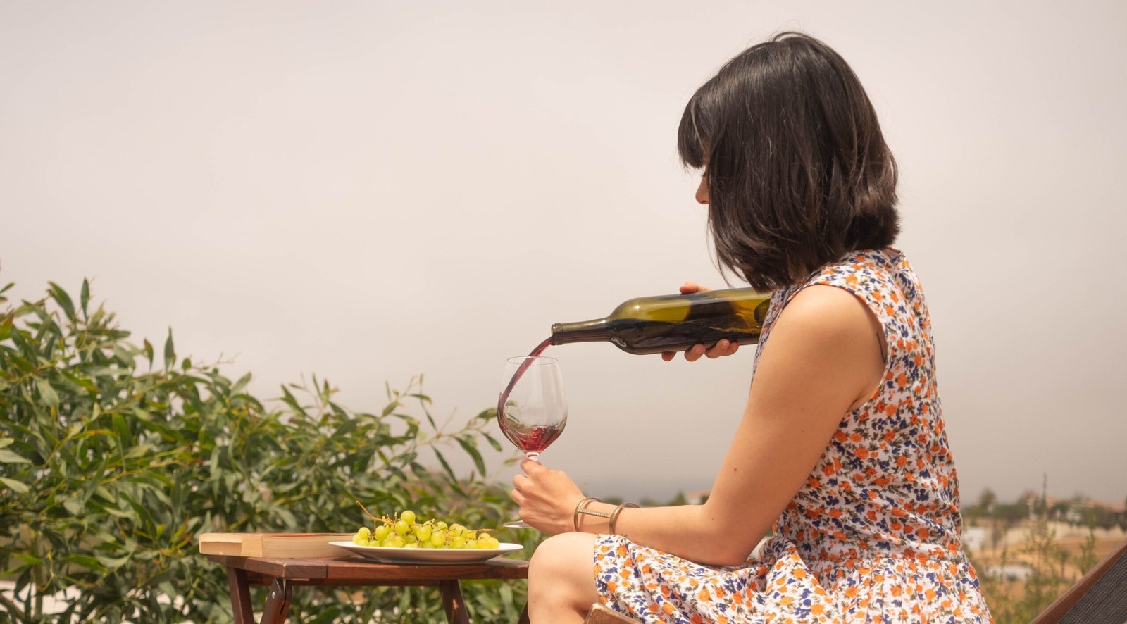 A woman wearing a floral dress pours wine into a glass while sitting outdoors on a sunny day.